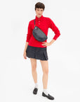 A model wearing a vibrant red long-sleeve polo shirt, a dark gray pleated mini skirt, white socks, and black loafers, accessorized with a dark navy woven crossbody bag.