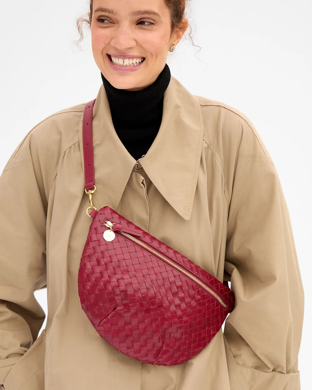 A woman wearing a beige trench coat over a black turtleneck, carrying a burgundy woven leather crescent-shaped crossbody bag with gold hardware.