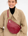 A woman wearing a beige trench coat over a black turtleneck, carrying a burgundy woven leather crescent-shaped crossbody bag with gold hardware.