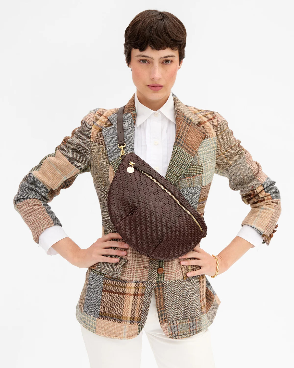 A model wearing a brown woven crossbody bag with gold hardware over a multicolor patchwork blazer and white shirt, against a plain white background.