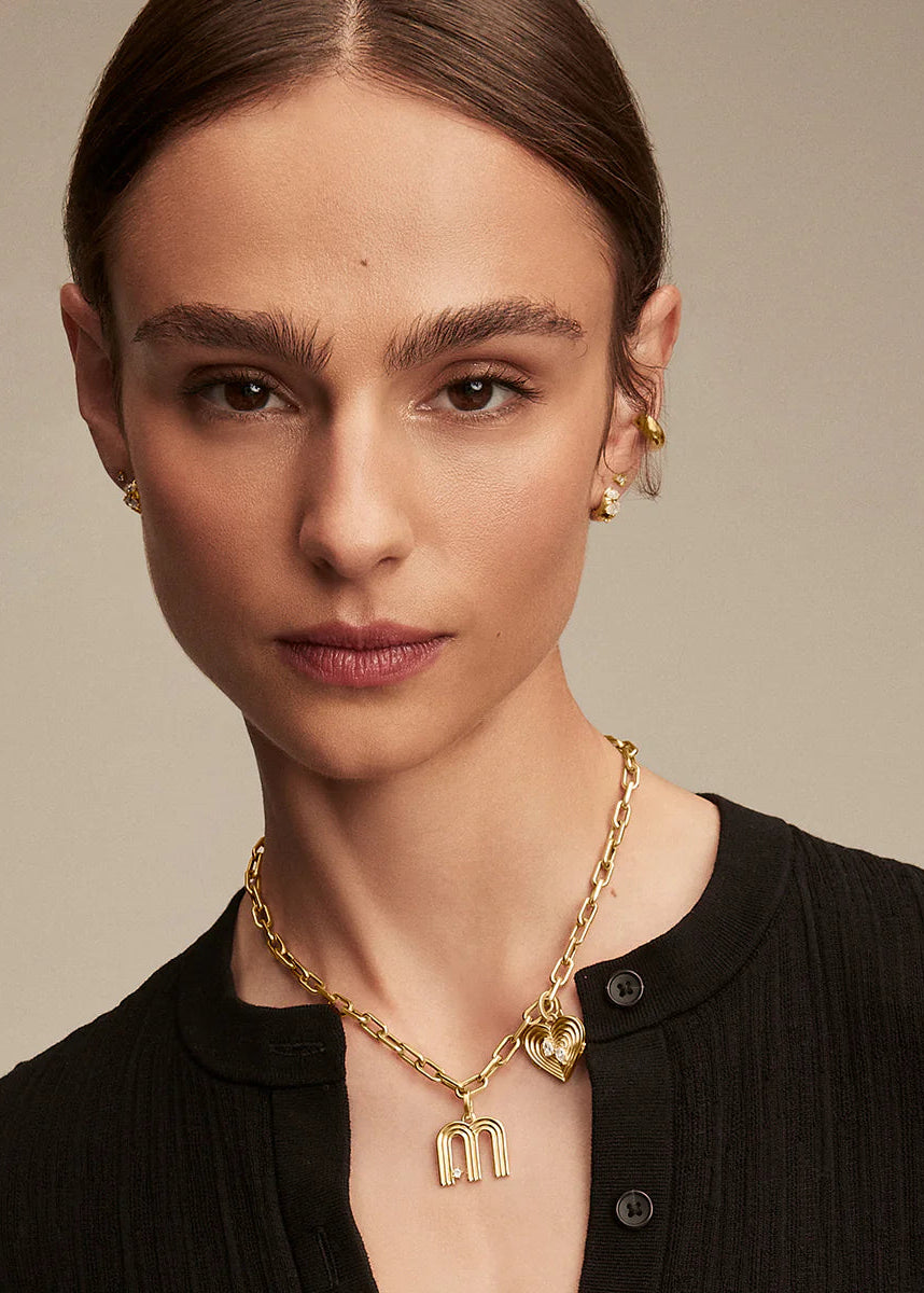 Woman wearing a gold necklace with a 'M' pendant against a neutral background