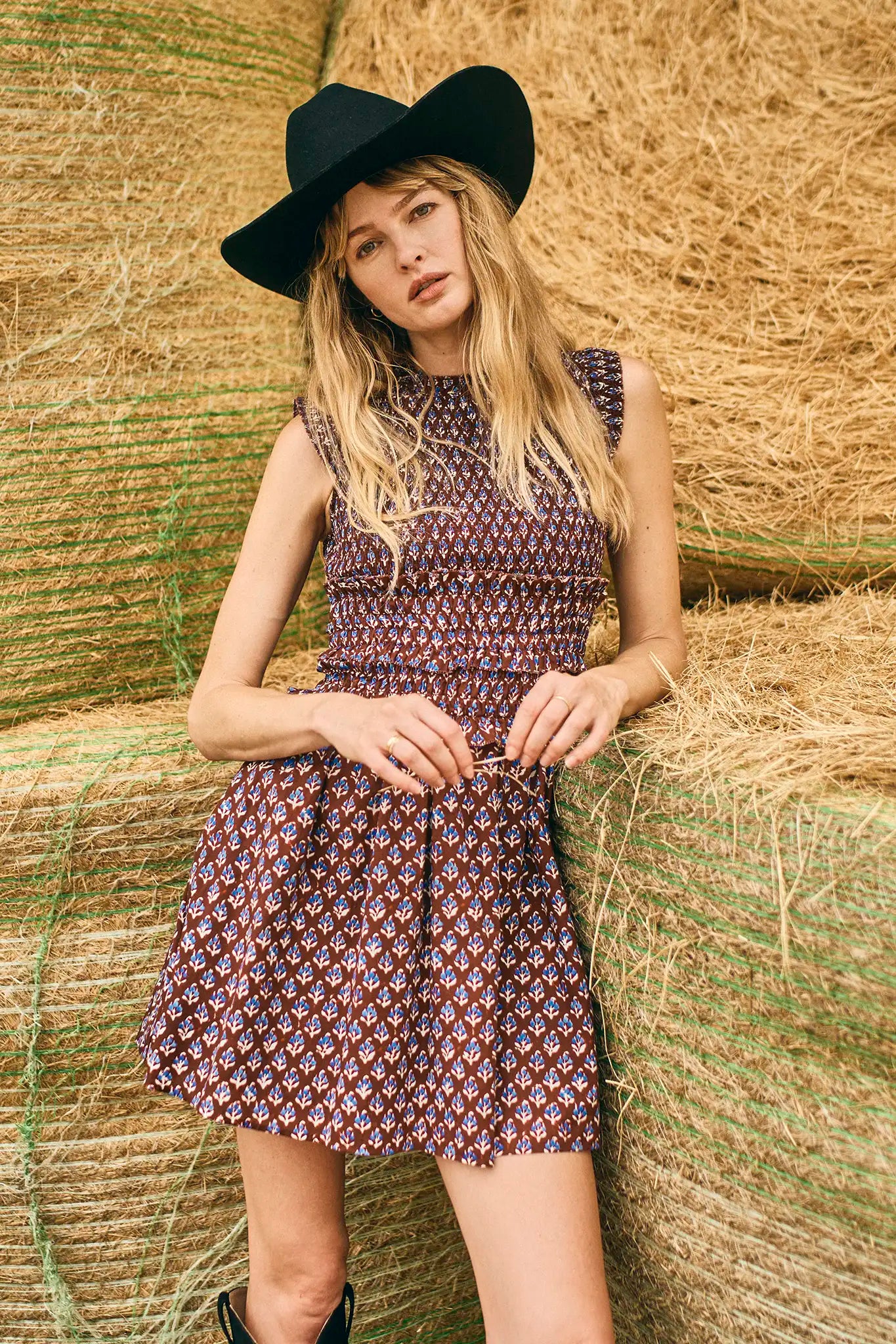 Woman in a patterned dress and black hat standing among hay bales.