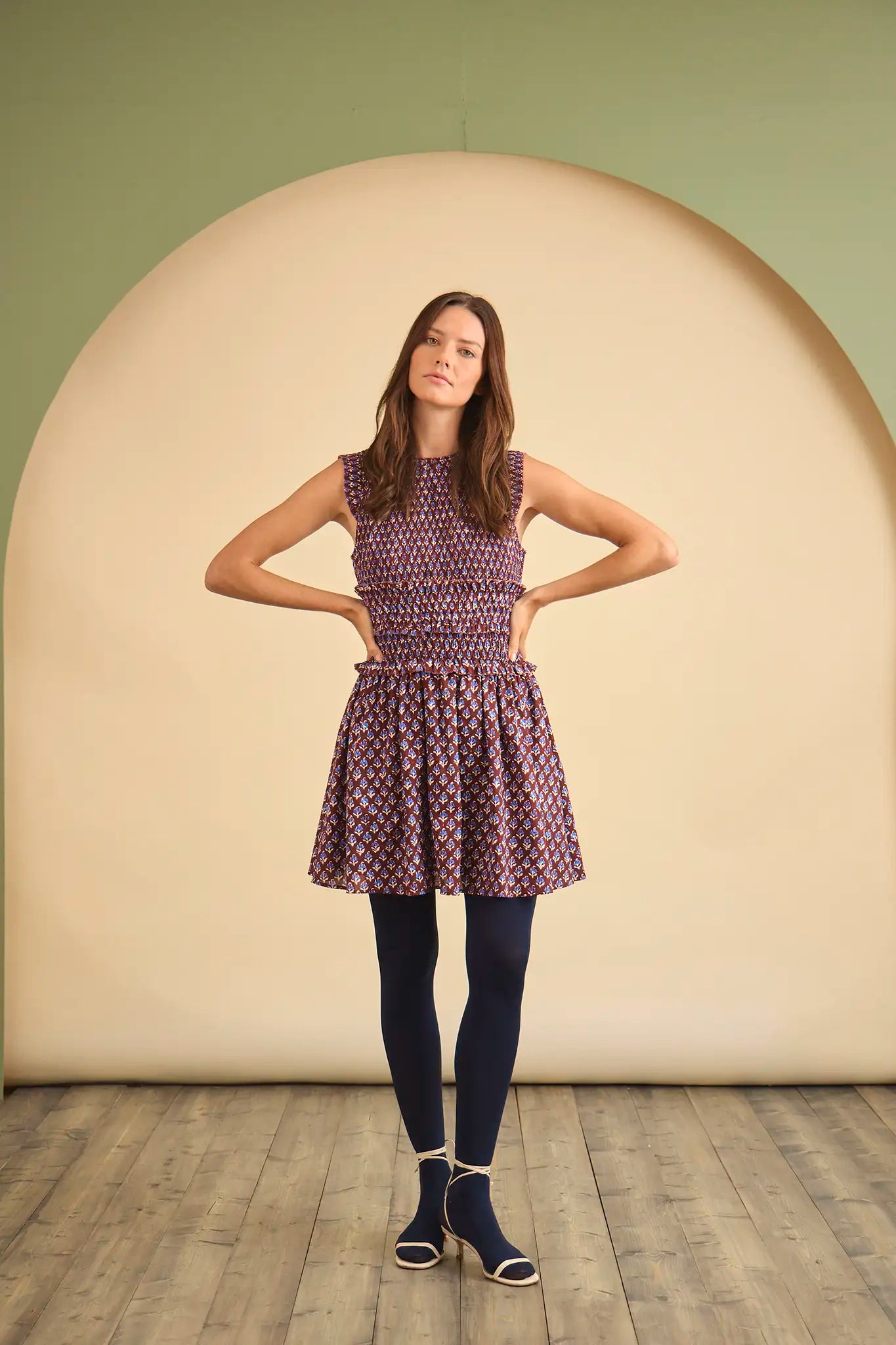 Woman wearing a patterned dress and dark tights standing against a plain background