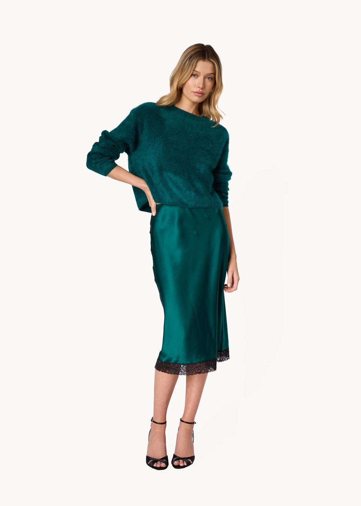 A woman modeling a dark teal green two-piece set consisting of a textured long-sleeve top and a satin midi skirt with a black lace hem, paired with black strappy heels against a plain white background.