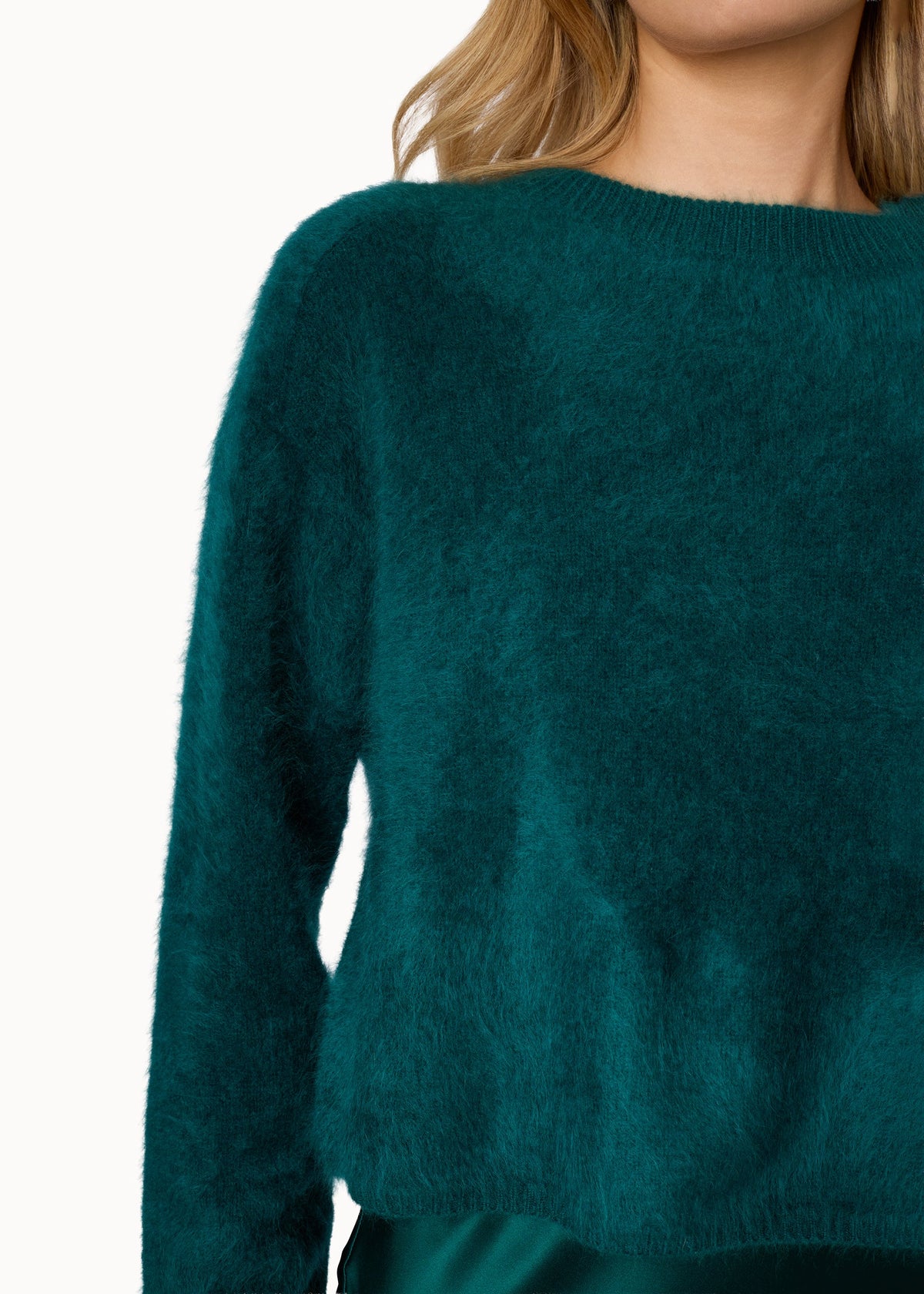 Close-up of a model wearing a fuzzy, long-sleeve knit sweater in a deep teal green color, paired with a matching satin skirt against a plain white background.