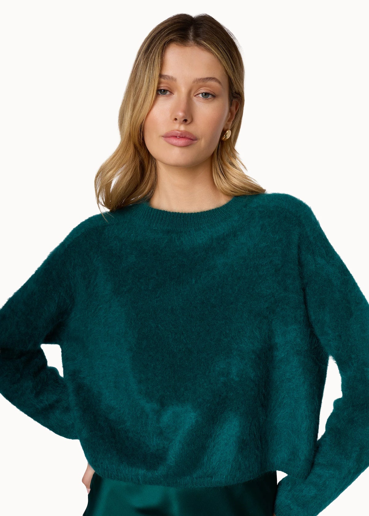 A woman wearing a fuzzy, cropped long-sleeve sweater in a rich teal green color, paired with a matching satin skirt, against a plain white studio background.