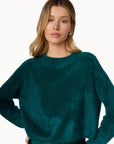 A woman wearing a fuzzy, cropped long-sleeve sweater in a rich teal green color, paired with a matching satin skirt, against a plain white studio background.