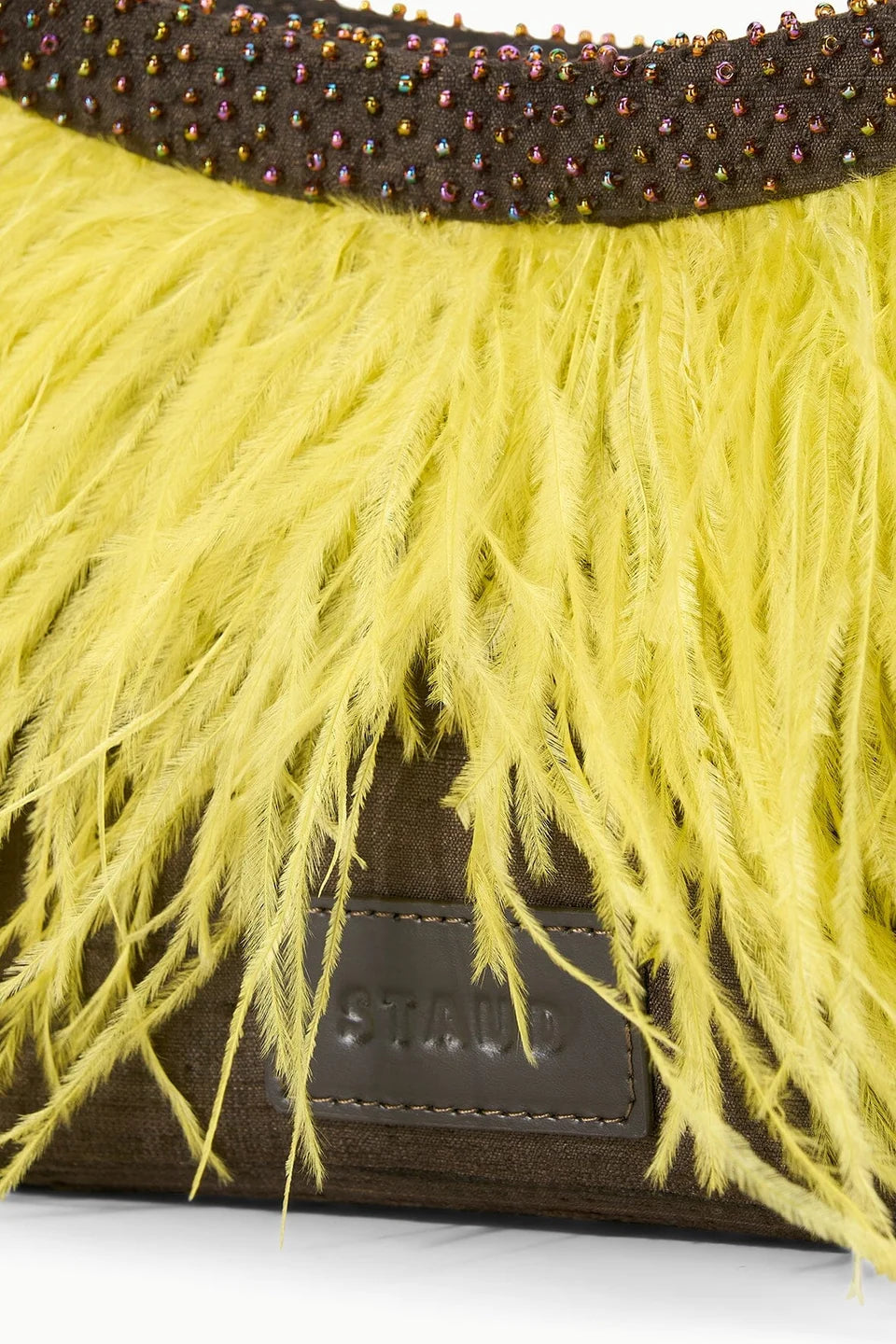Close-up of a Staud handbag featuring bright yellow ostrich feather fringe over a dark brown suede body with a beaded waistband and a Staud leather logo patch.