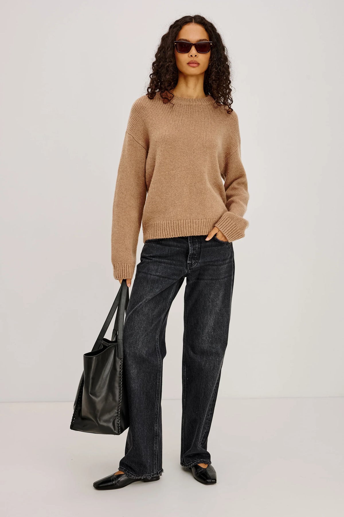 Woman wearing a beige sweater and black jeans, holding a black bag against a white background
