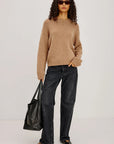 Woman wearing a beige sweater and black jeans, holding a black bag against a white background