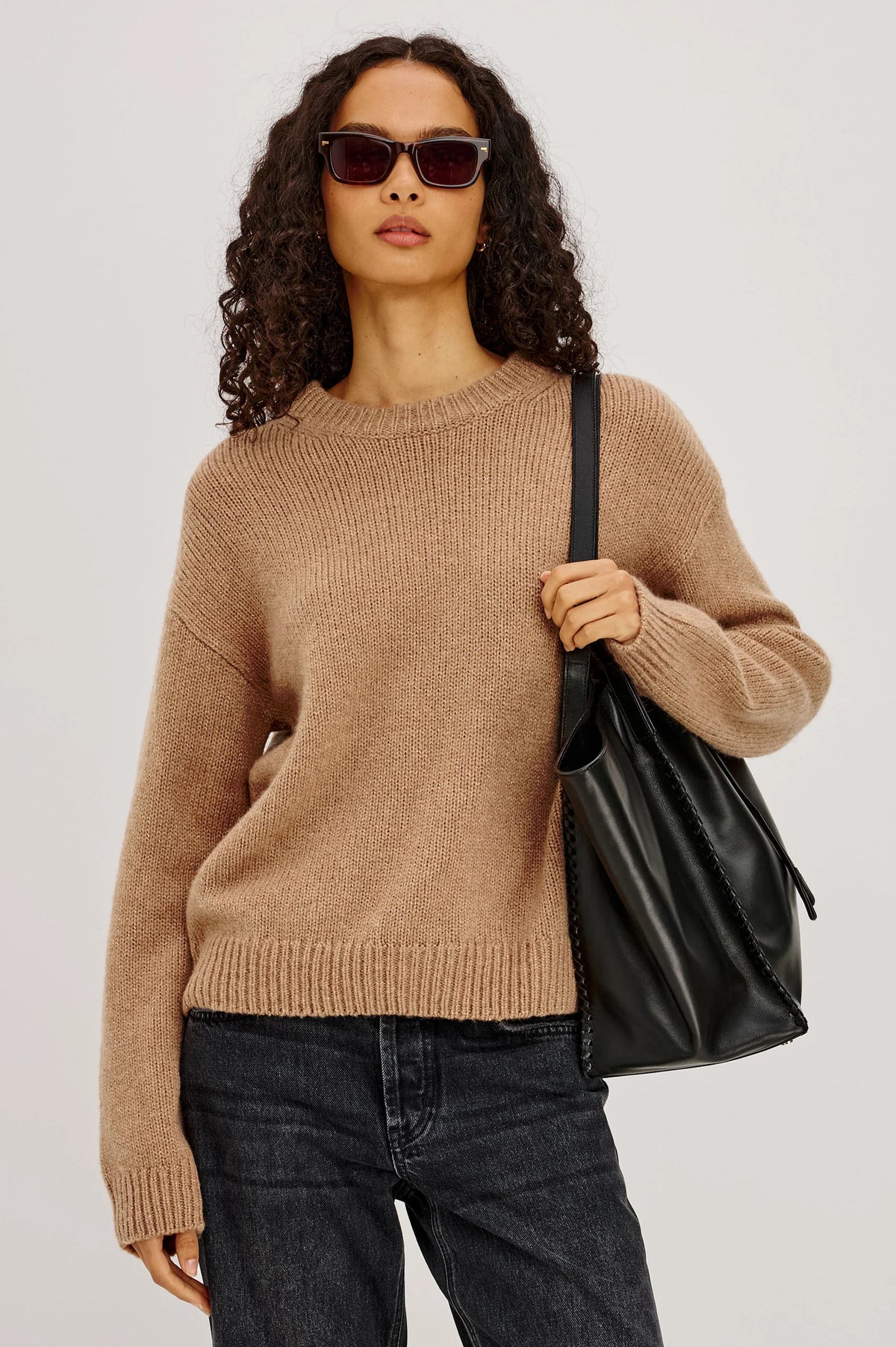 Woman wearing a beige sweater and dark jeans, holding a black bag against a plain background