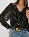 Woman wearing a black polka dot blouse with sheer sleeves and blue jeans on a light gray background