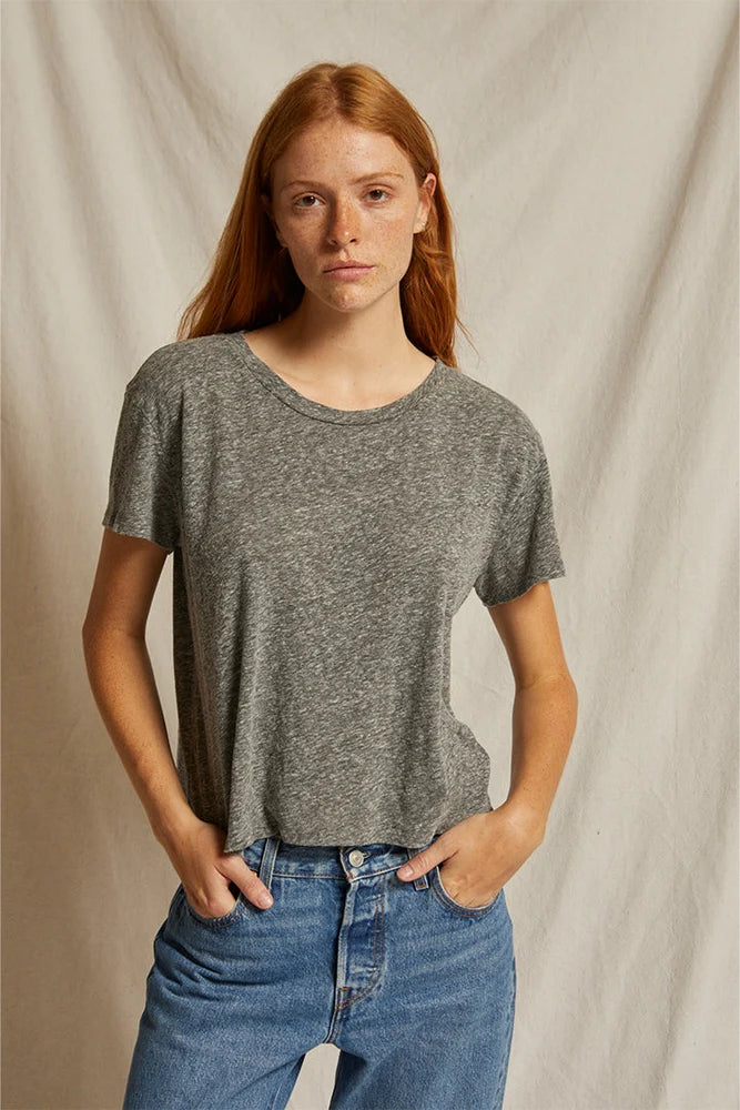 A woman with red hair wearing a heather gray short-sleeve crewneck t-shirt and blue jeans, standing against a draped beige fabric backdrop.