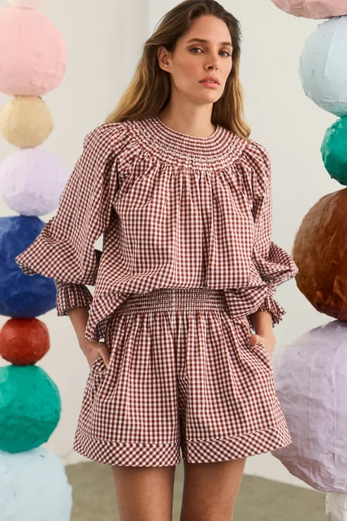A woman wearing a red and white gingham smocked set with ruffled sleeves, standing in front of a white wall decorated with colorful paper lanterns.