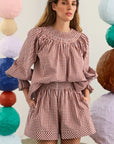 A woman wearing a red and white gingham smocked set with ruffled sleeves, standing in front of a white wall decorated with colorful paper lanterns.