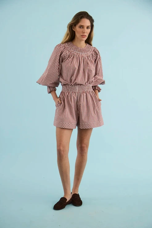 A woman modeling a pink and white gingham two-piece set consisting of a long-sleeve blouse with ruffled sleeves and matching shorts, paired with dark brown suede loafers, against a solid light blue studio background.