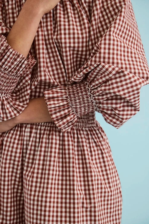 A close-up of a person wearing a red and white gingham dress with voluminous ruffled sleeves and a smocked waist, set against a solid light blue background.
