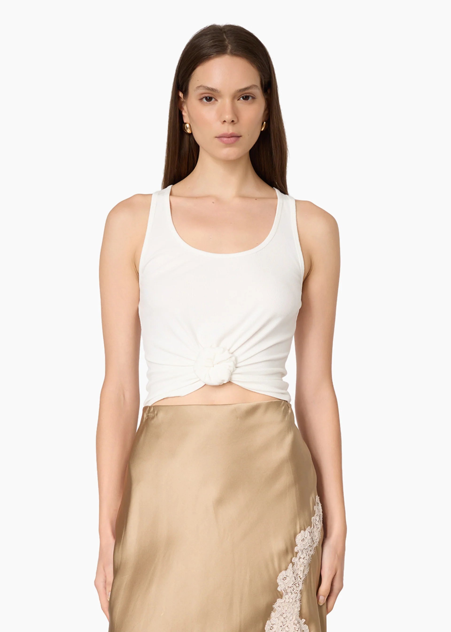 A model wearing a white ribbed knit tank top with a twisted front detail and a beige satin skirt with lace trim, set against a plain light gray background.