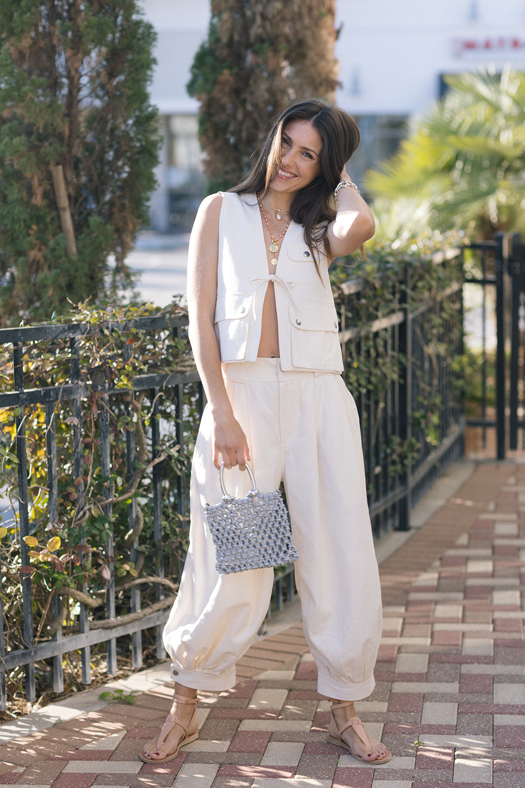 Woman in a white outfit standing on a sidewalk with greenery in the background