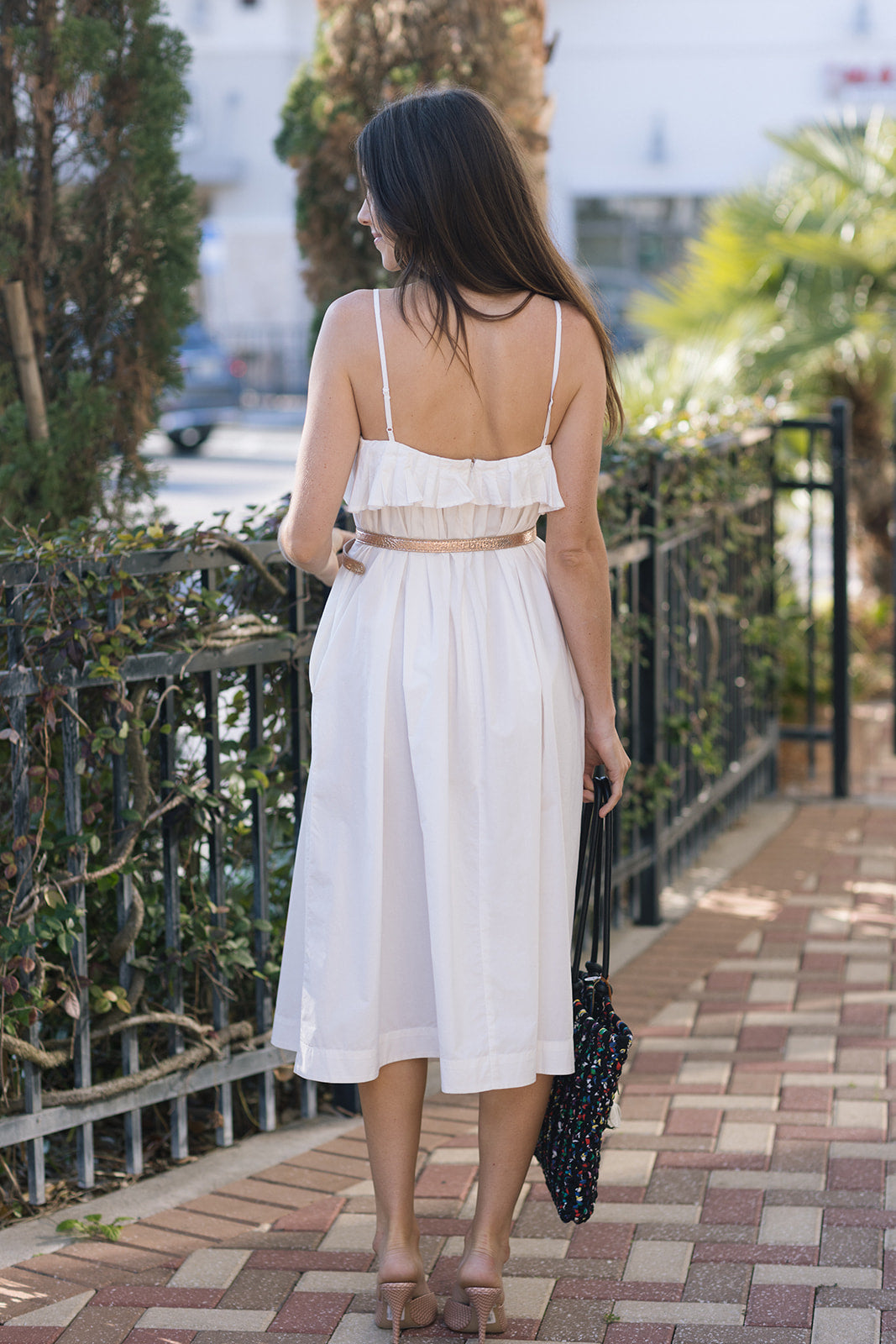 Woman in a white dress walking on a sidewalk with greenery and buildings in the background