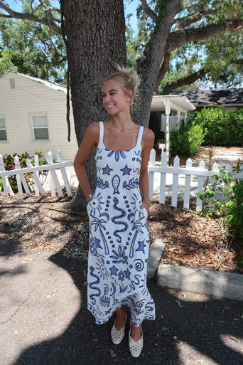 Woman in a white dress with blue patterns standing outdoors near a tree and house.