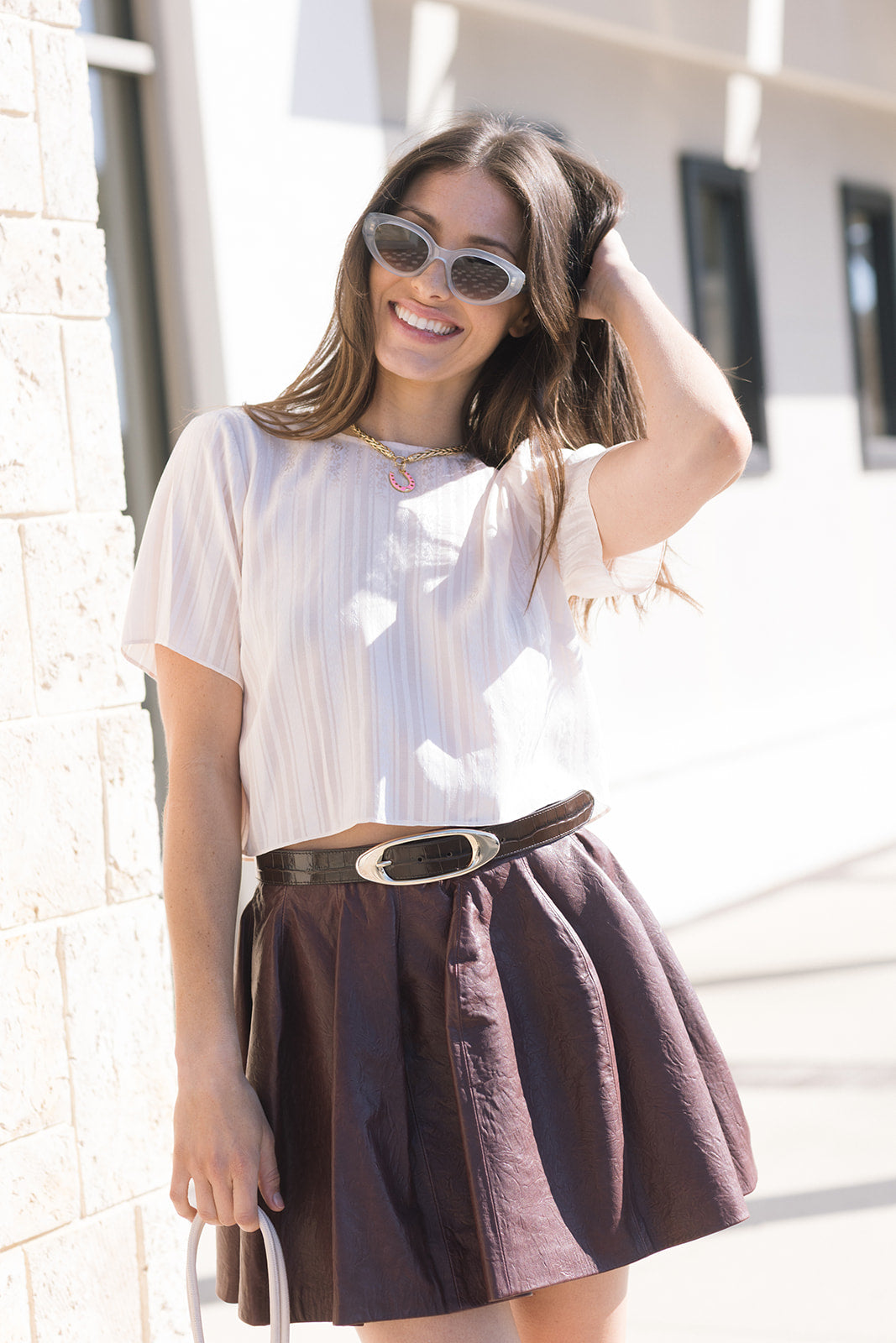 Woman wearing a white blouse and brown skirt with Candice Croco Belt and sunglasses, standing against a light-colored wall.