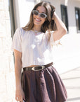 Woman wearing a white blouse and brown skirt with Candice Croco Belt and sunglasses, standing against a light-colored wall.