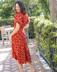 A woman wearing a red and orange patterned midi dress with short puff sleeves, standing on a brick patio in an outdoor garden setting.