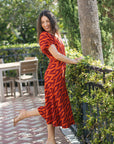 A woman posing outdoors in a red and black patterned midi dress with beige high heels, leaning on a black metal fence next to a green hedge.