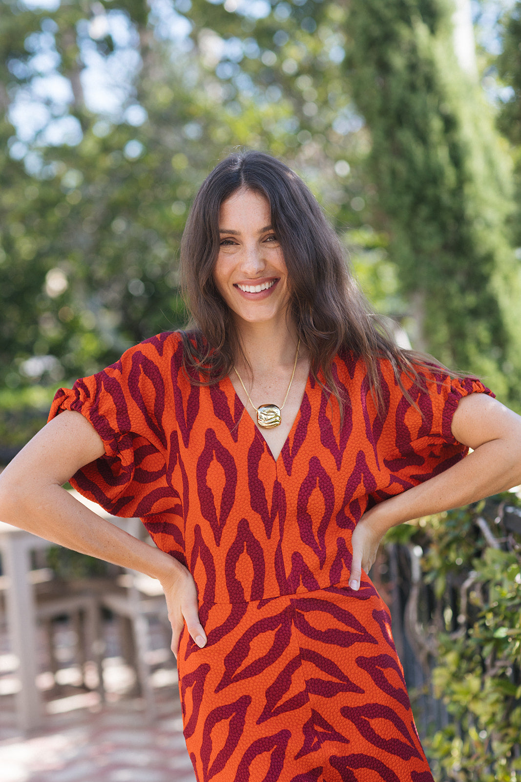 A woman wearing a short-sleeve V-neck dress with an orange and dark red abstract pattern, standing outdoors with her hands on her hips against a blurred green background.