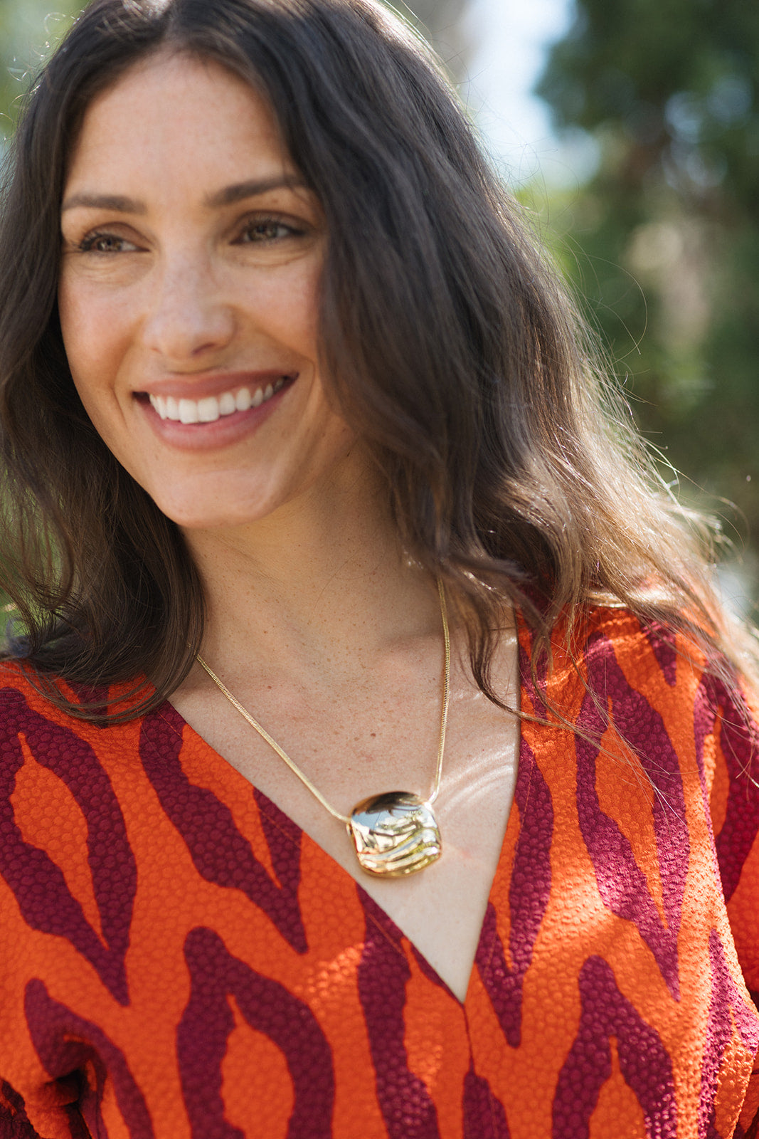 A smiling woman with long dark hair wearing a textured orange and burgundy patterned top and a gold pendant necklace, photographed outdoors with a blurred green background.