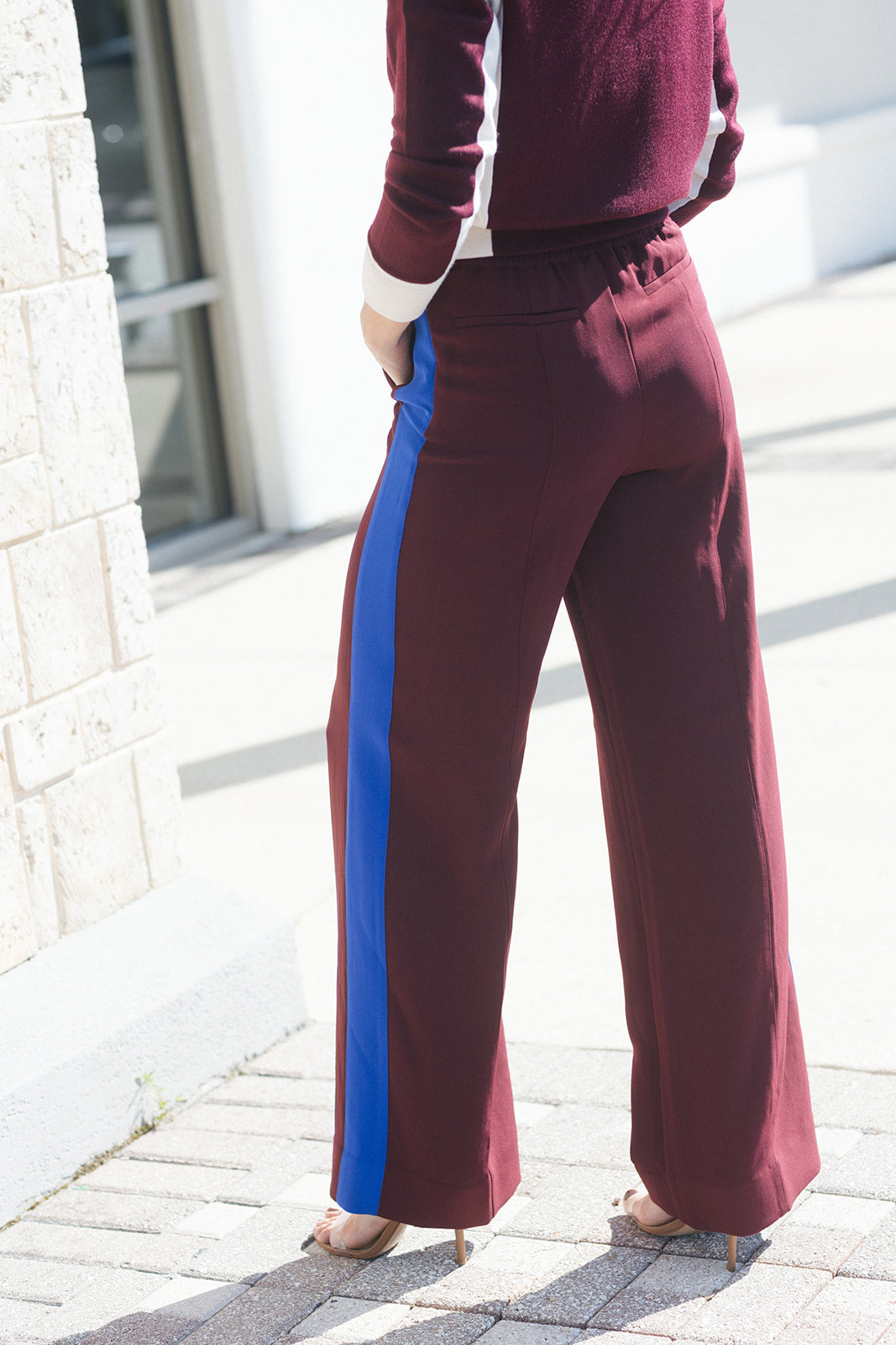 A person wearing wide-leg maroon pants with a bold blue side stripe, paired with a matching maroon top and beige heels, standing outdoors on a paved walkway.
