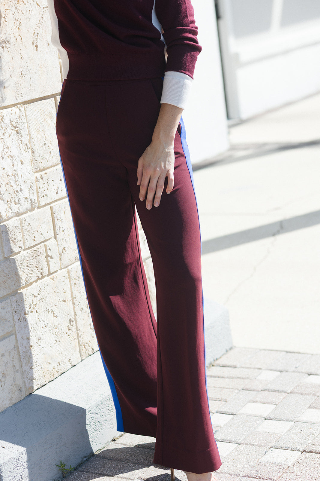 A person wearing burgundy wide-leg pants with blue side panels and a matching burgundy top, standing outdoors against a light stone wall and paved ground.