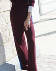 A person wearing burgundy wide-leg pants with blue side panels and a matching burgundy top, standing outdoors against a light stone wall and paved ground.