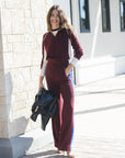 model wearing a burgundy sweater with white details and burgundy pants with a blue stripe next to a brick wall in a shopping plaza 