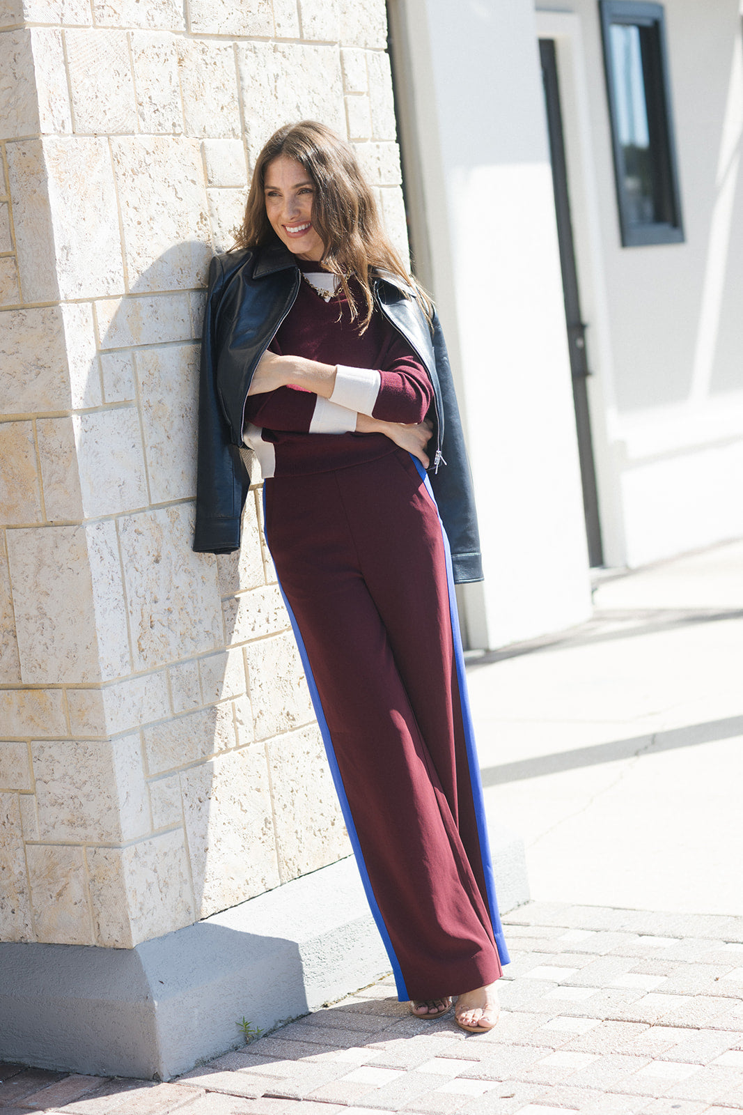model wearing burgundy pants with a blue stripe wearing leather jacket and a burgundy sweater with white details with a shopping center as the background