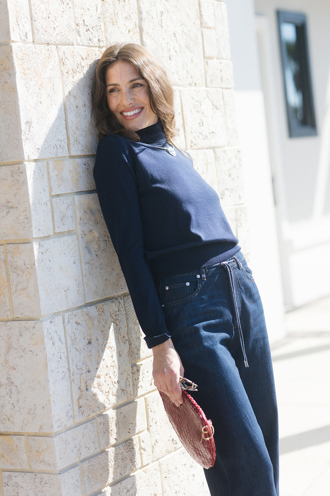 A woman wearing a navy blue mock neck top and matching dark blue jeans leans against a light beige stone wall, holding a textured red clutch.