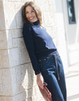 A woman wearing a navy blue mock neck top and matching dark blue jeans leans against a light beige stone wall, holding a textured red clutch.
