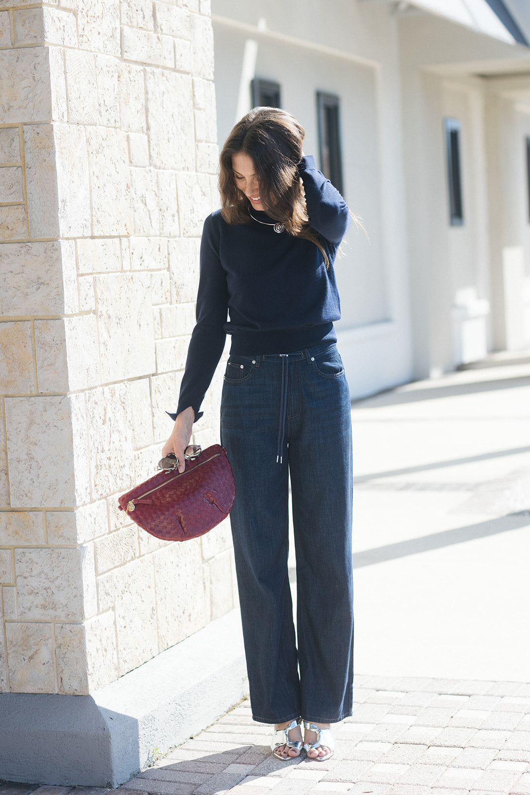 A woman stands outdoors in a sunny alleyway wearing a navy blue long-sleeve top, dark wide-leg denim jeans, and holding a burgundy textured handbag.
