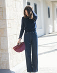A woman stands outdoors in a sunny alleyway wearing a navy blue long-sleeve top, dark wide-leg denim jeans, and holding a burgundy textured handbag.
