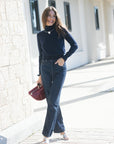A woman stands outdoors on a paved walkway wearing a dark navy long-sleeve mock neck top and dark blue flare jeans, holding a burgundy handbag.