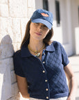 Woman wearing a blue cap with red lips and navy quilted vest leaning against a stone wall.