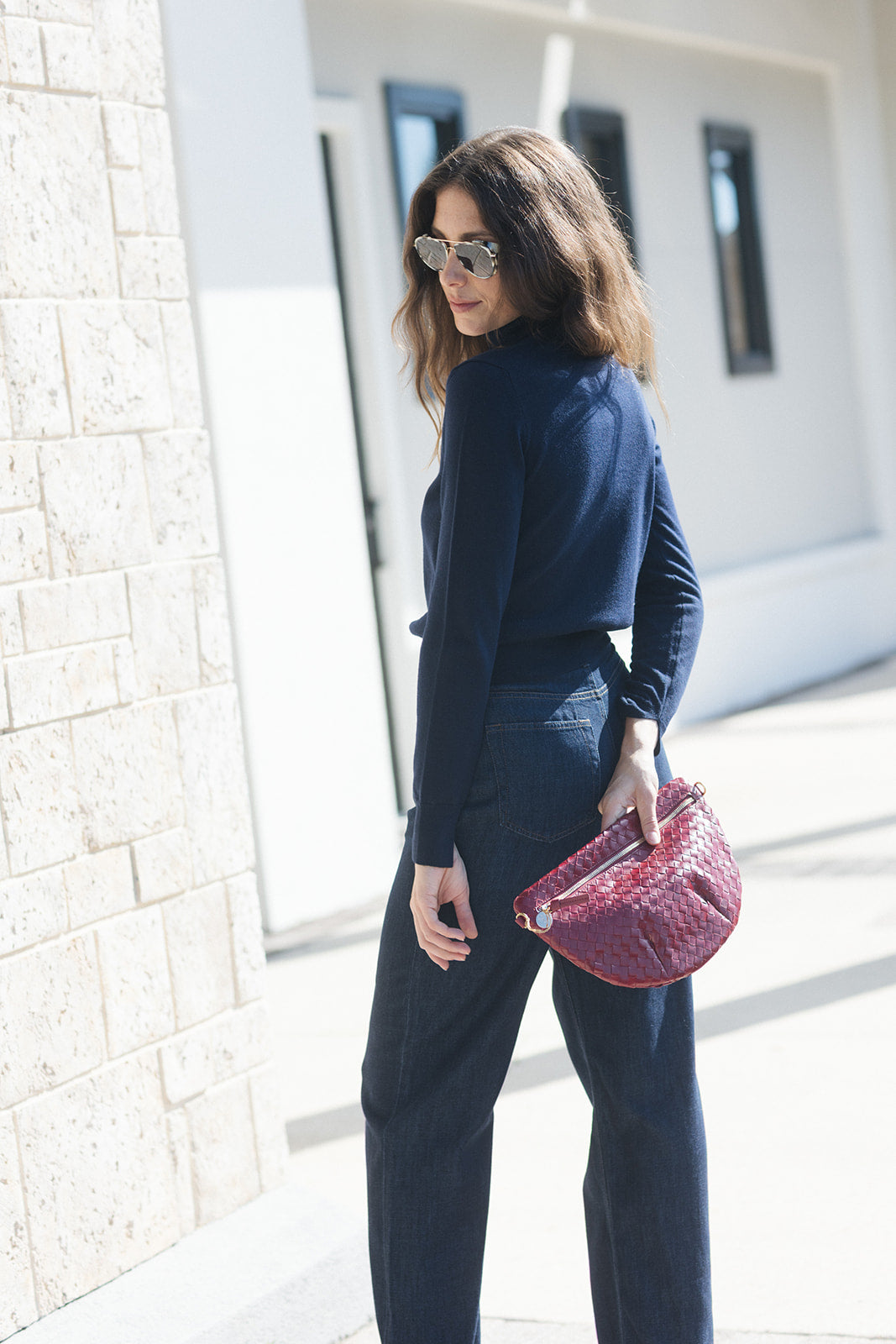 A woman wearing a navy blue long-sleeve crop top and dark blue jeans, holding a textured pink clutch bag, standing outdoors next to a light stone wall.
