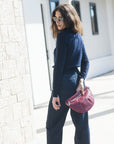 A woman wearing a navy blue long-sleeve crop top and dark blue jeans, holding a textured pink clutch bag, standing outdoors next to a light stone wall.