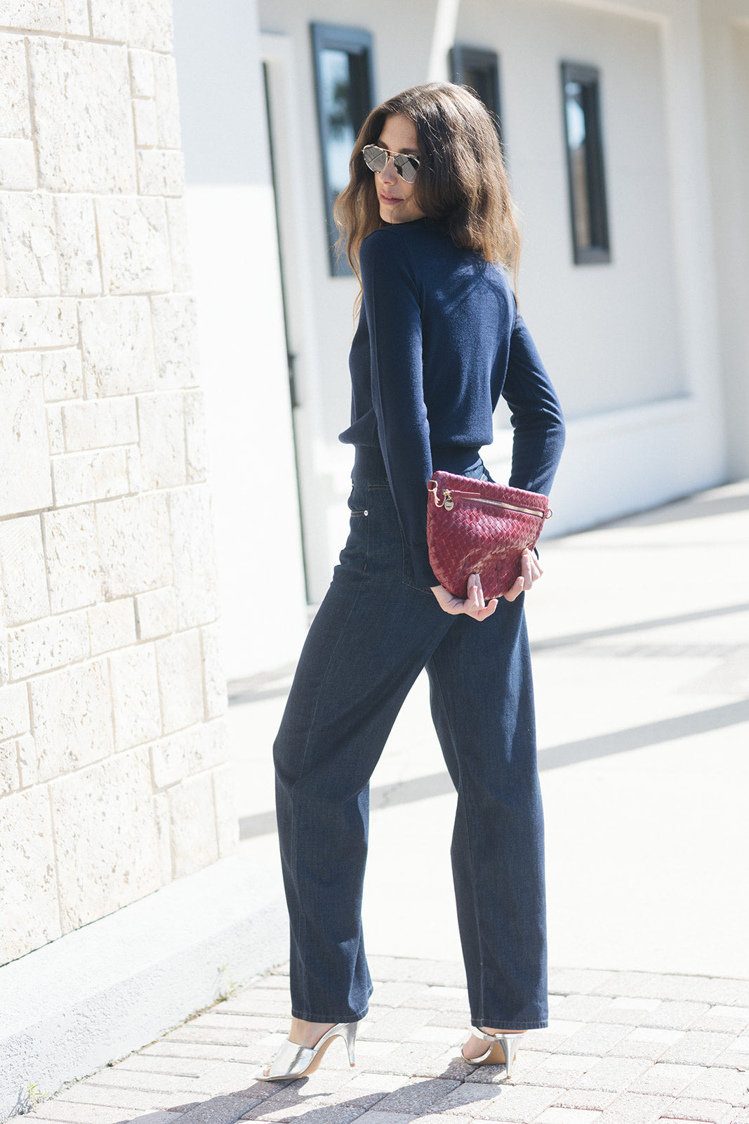 A woman wearing a navy blue long-sleeve top and matching wide-leg pants, holding a textured burgundy clutch bag, standing outdoors next to a light-colored stone wall.