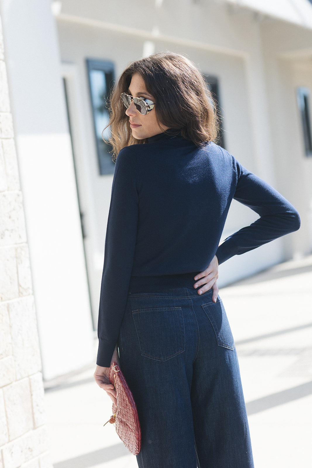 A woman wearing a navy blue mock neck long-sleeve top and dark blue wide-leg jeans, holding a pink patterned clutch bag, photographed outdoors against a bright white building.
