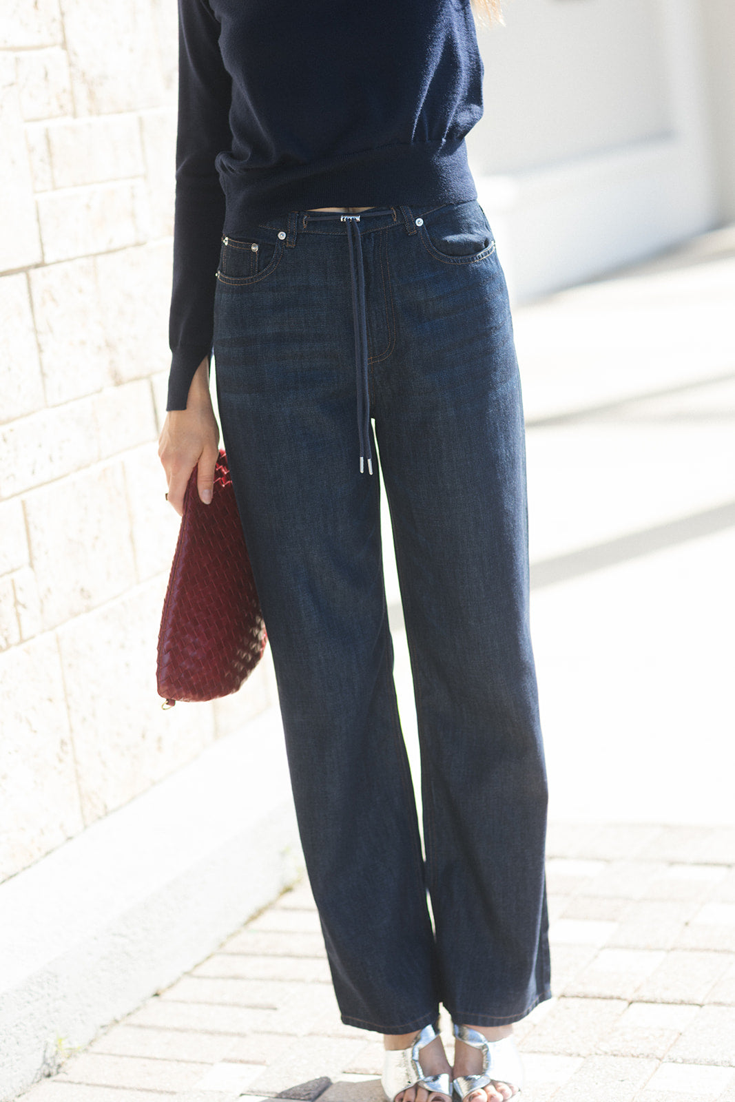 A person wearing dark blue wide-leg jeans with a drawstring waist, paired with a navy long-sleeve top and holding a burgundy woven clutch, standing outdoors against a light stone wall.
