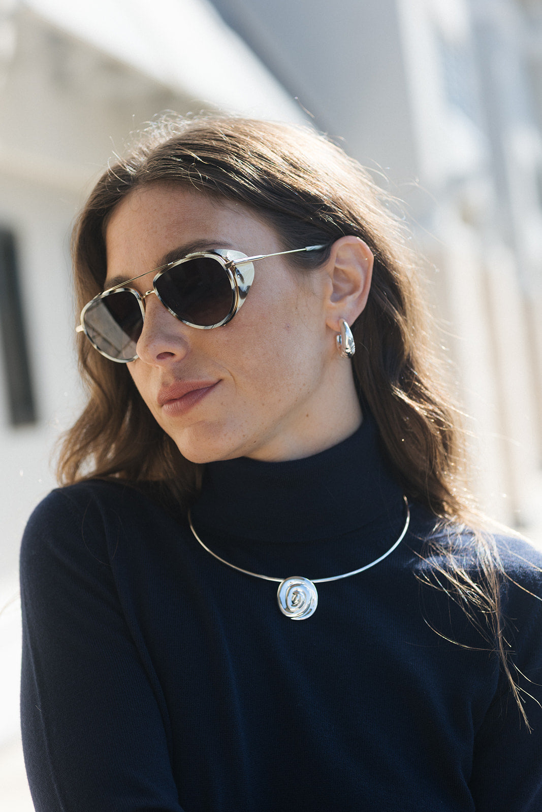 A woman wearing round silver-framed sunglasses, a silver pendant necklace with a circular logo, and a small hoop earring, dressed in a dark navy turtleneck sweater.