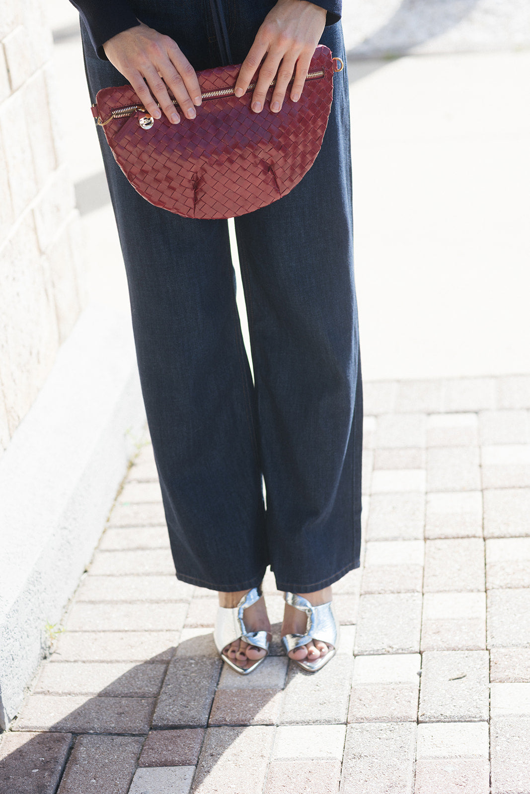 A person outdoors wearing dark wide-leg pants, a dark top, and metallic silver heeled sandals, holding a burgundy woven clutch bag.