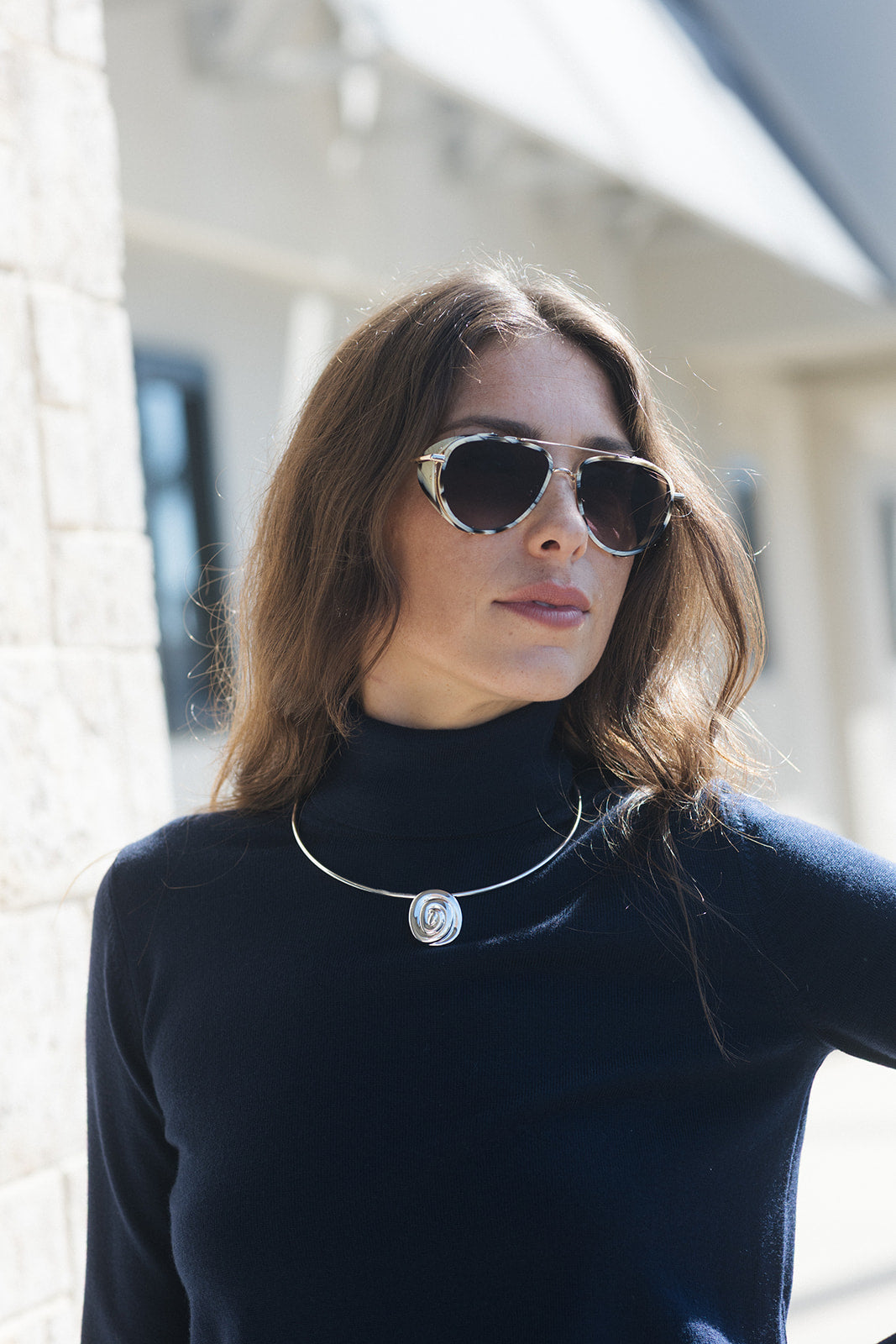A woman with brown hair wearing aviator sunglasses with a silver frame and a silver circular pendant necklace, dressed in a black turtleneck sweater, photographed outdoors against a blurred light-colored building background.